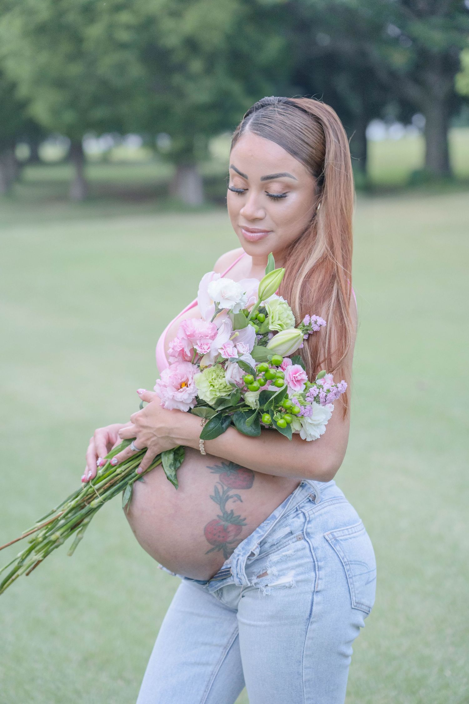 Pregnant woman with pink hair holding a bouquet of pink and white flowers, showing tattooed belly in a green park.