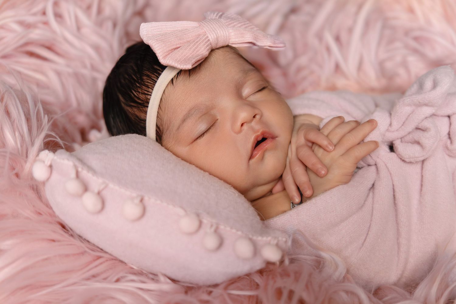 Peaceful newborn baby girl in pink wrap sleeping on a pom-pom pillow surrounded by pink fur.