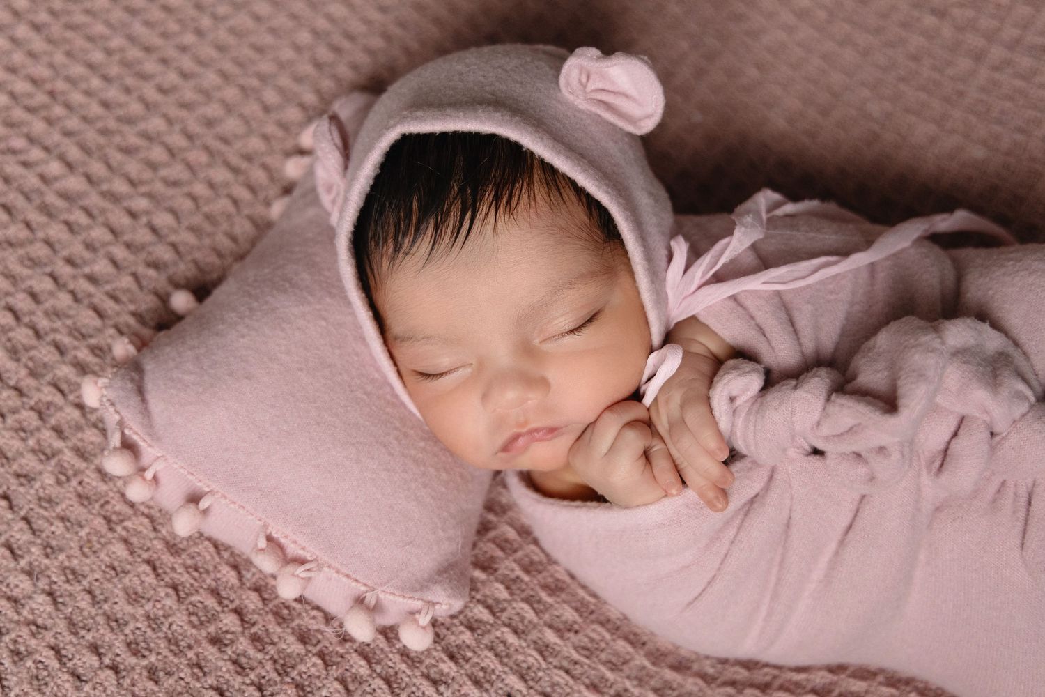 Sleeping newborn baby in pink bear bonnet and matching outfit on pink knit blanket with pillow.