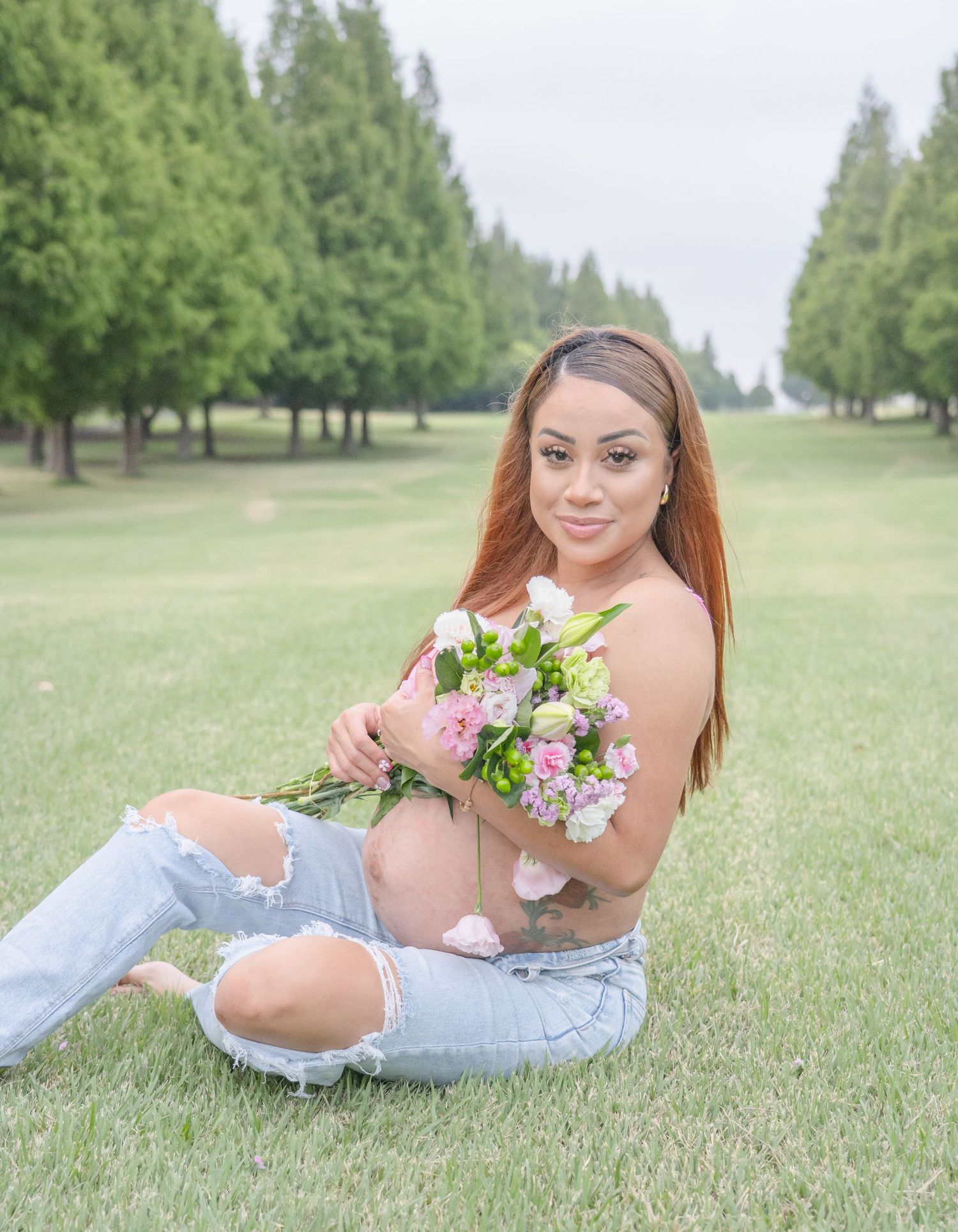 Pregnant woman with tattoos sitting on grass holding pink and white flowers in outdoor maternity photo.