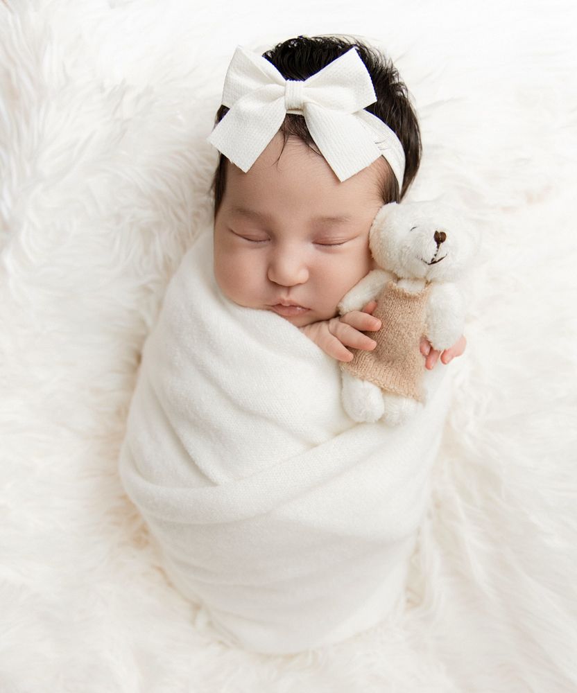 Sleeping newborn baby swaddled in white wrap holding teddy bear with white bow headband on fluffy white backdrop.