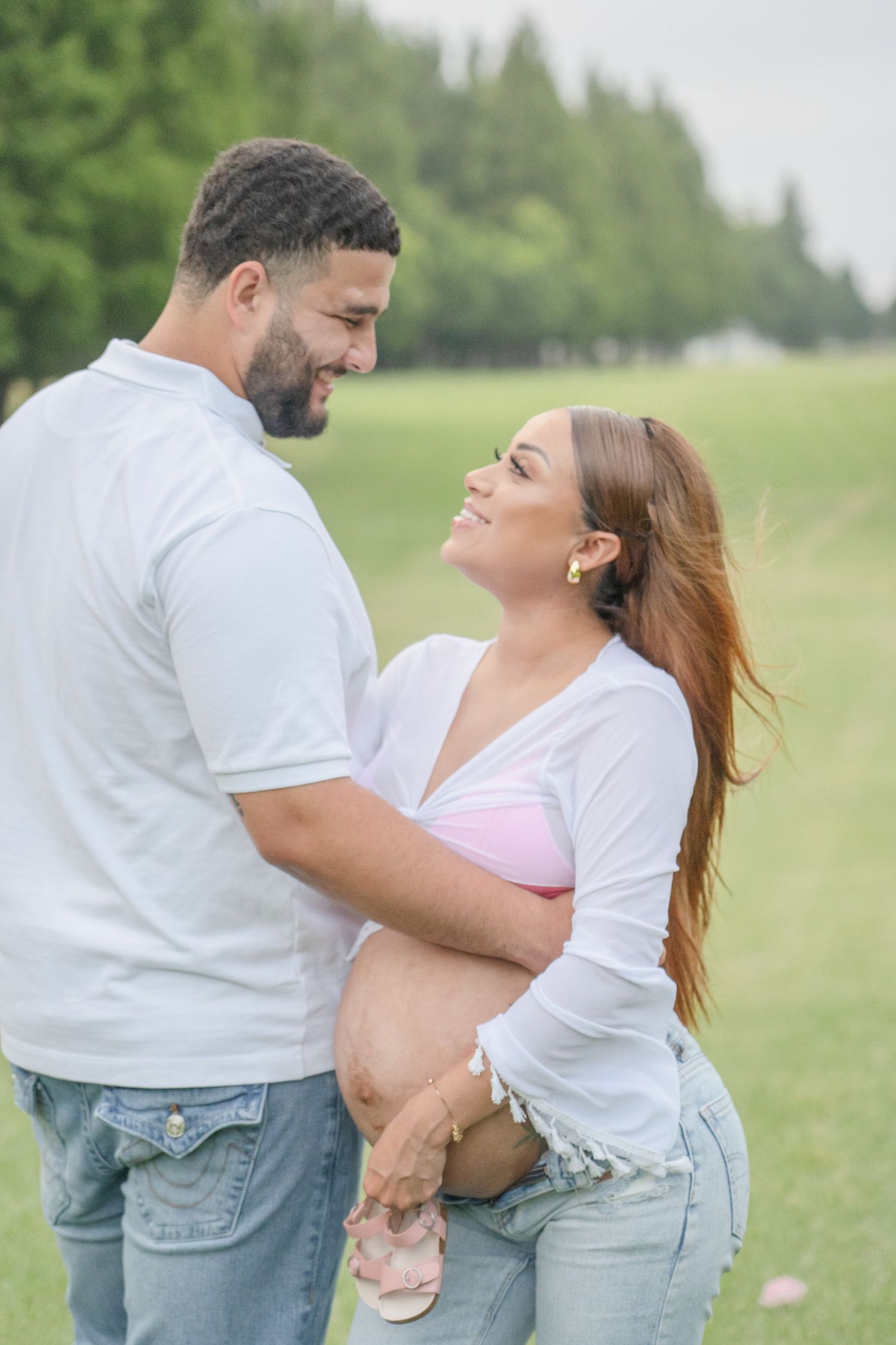 Smiling pregnant couple in white tops and jeans embracing outdoors on green grass during maternity photo session.