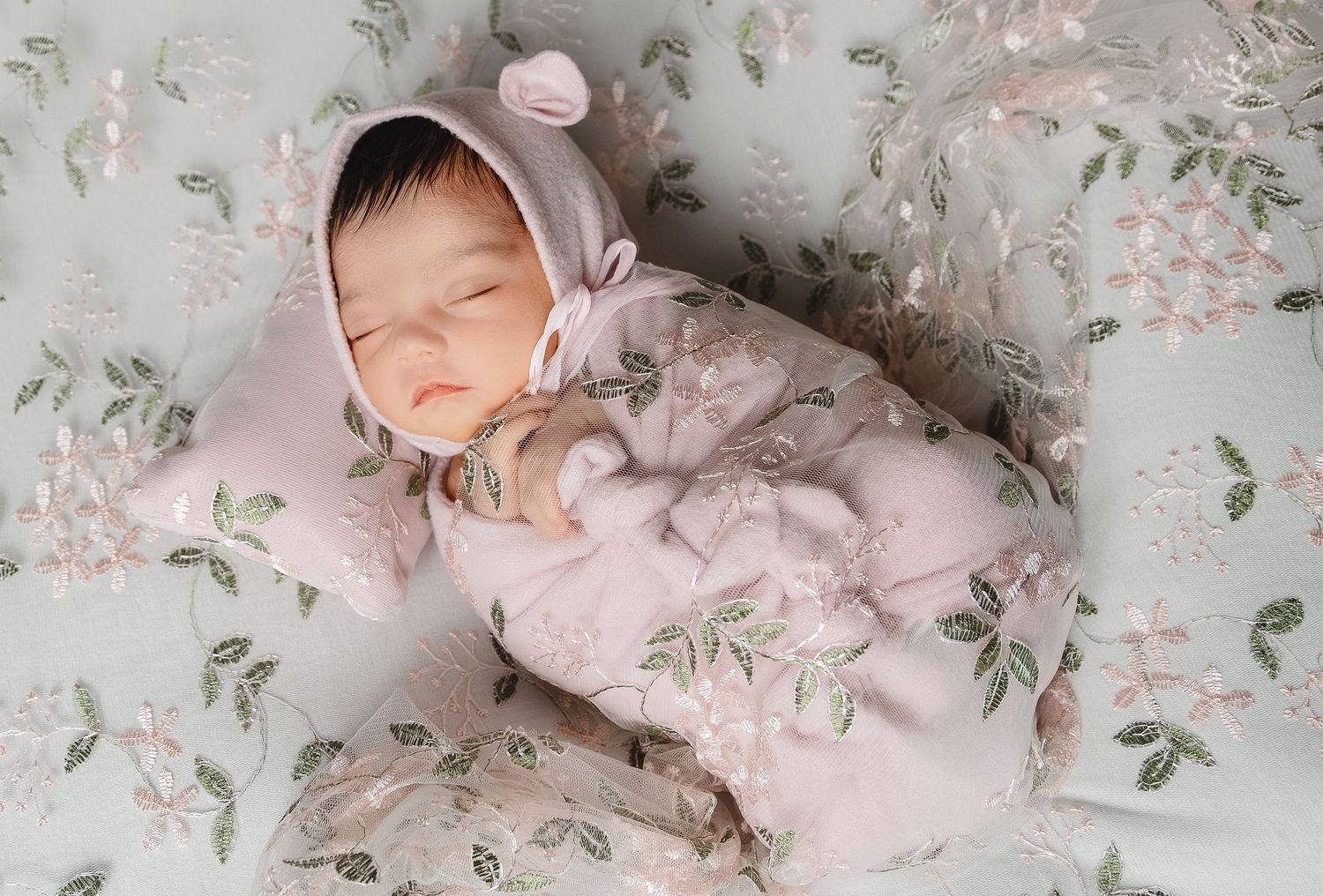 Sleeping newborn baby in pink bear bonnet outfit on floral embroidered blanket photography.