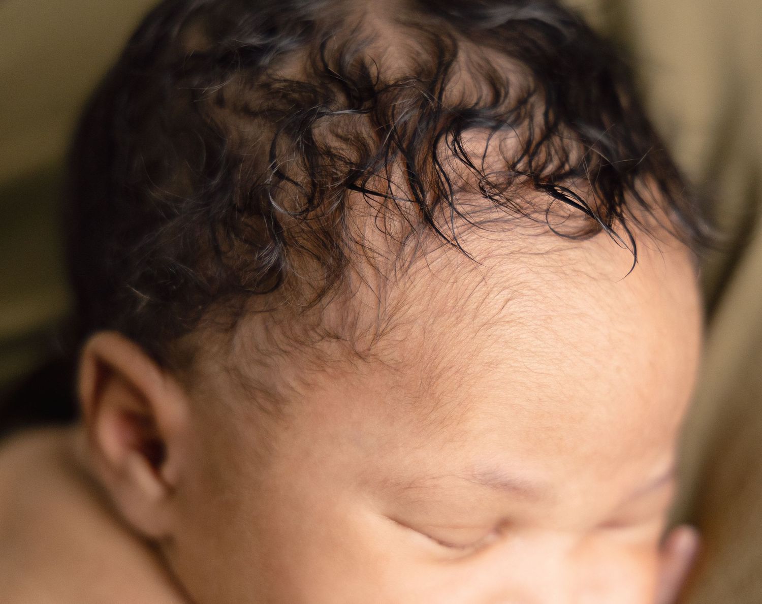Close-up of a sleeping newborn baby with dark curly hair against a warm olive background.