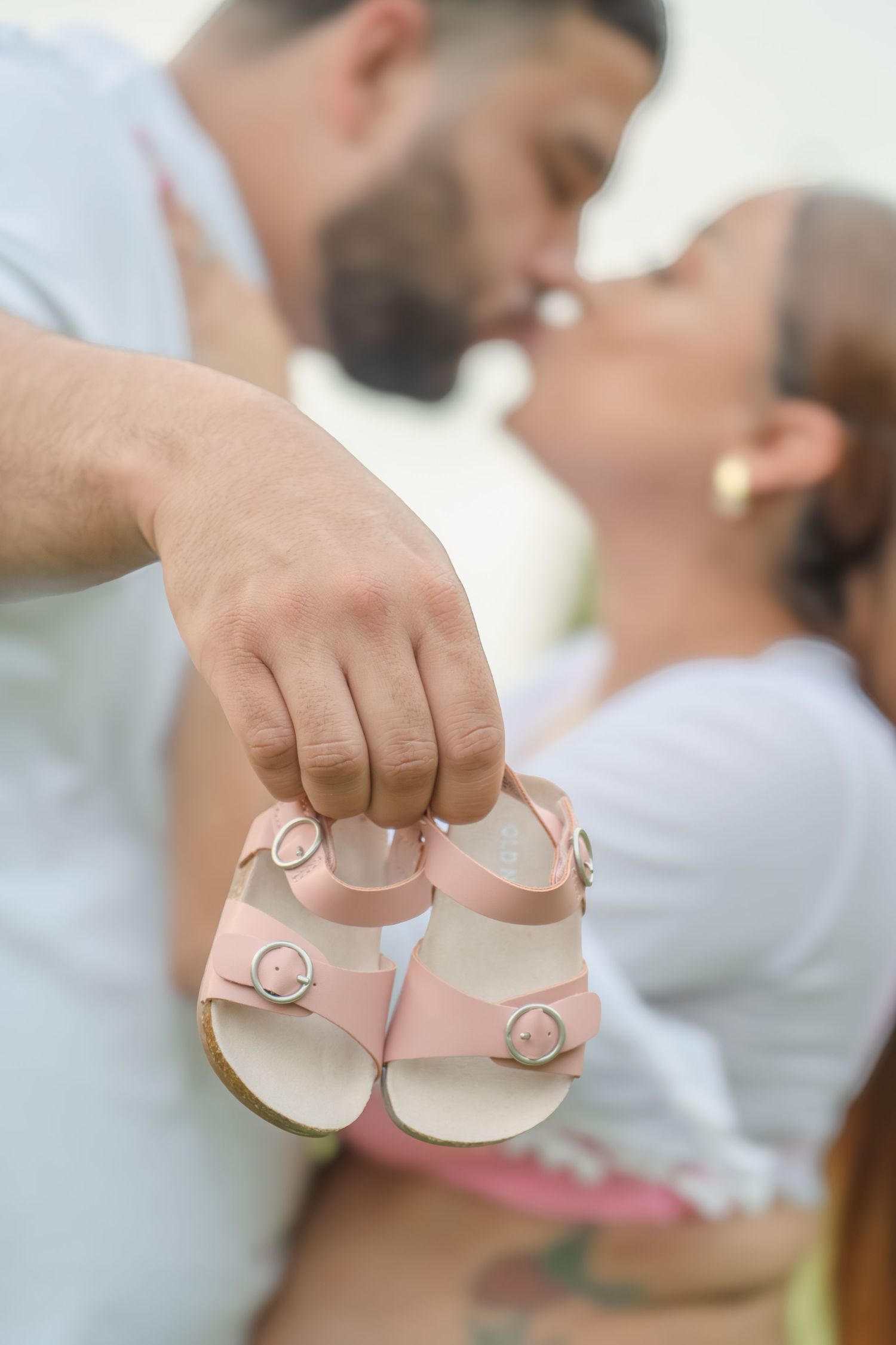 Couple kissing outdoors while man holds tiny pink baby sandals in foreground during maternity photo session.