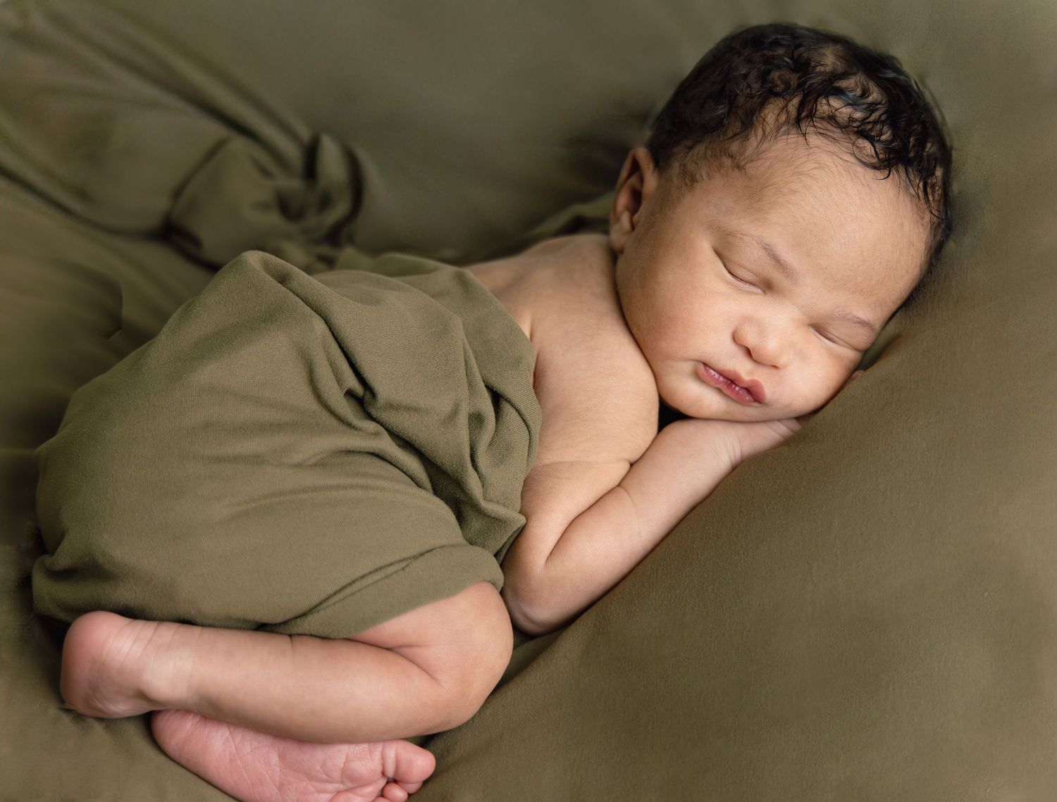 Newborn baby sleeping peacefully wrapped in olive green fabric on matching green background.