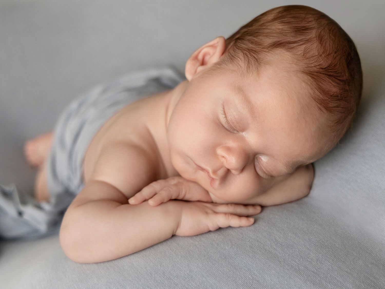 Newborn baby sleeping on grey blanket with hands tucked under cheek in studio photo.