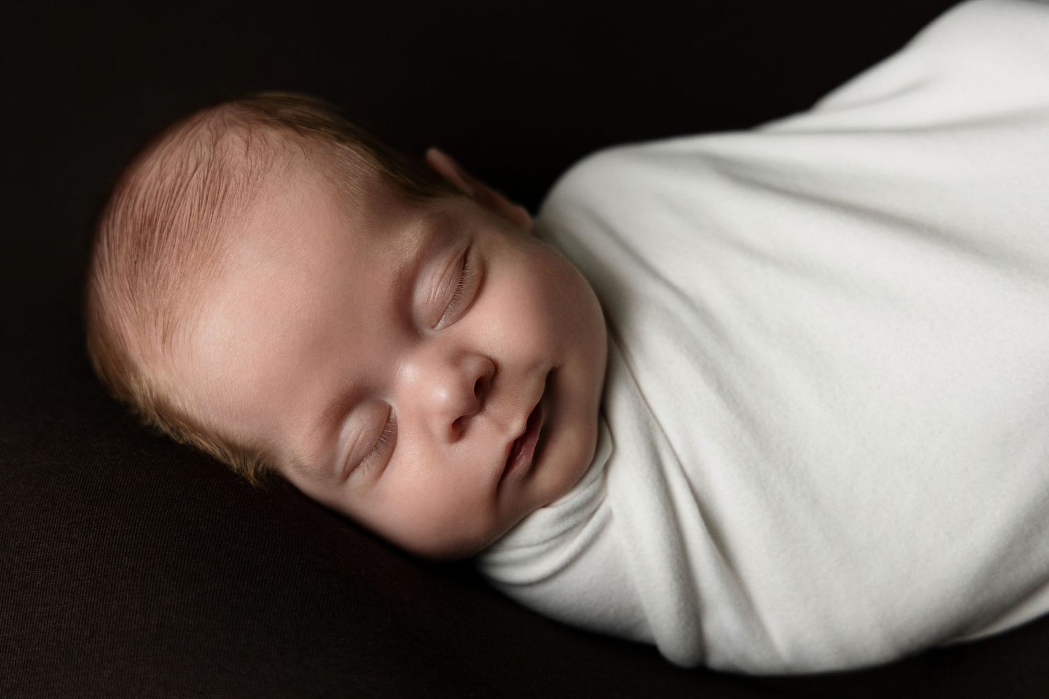 Peaceful newborn baby swaddled in white wrap sleeping on dark background in professional photo.