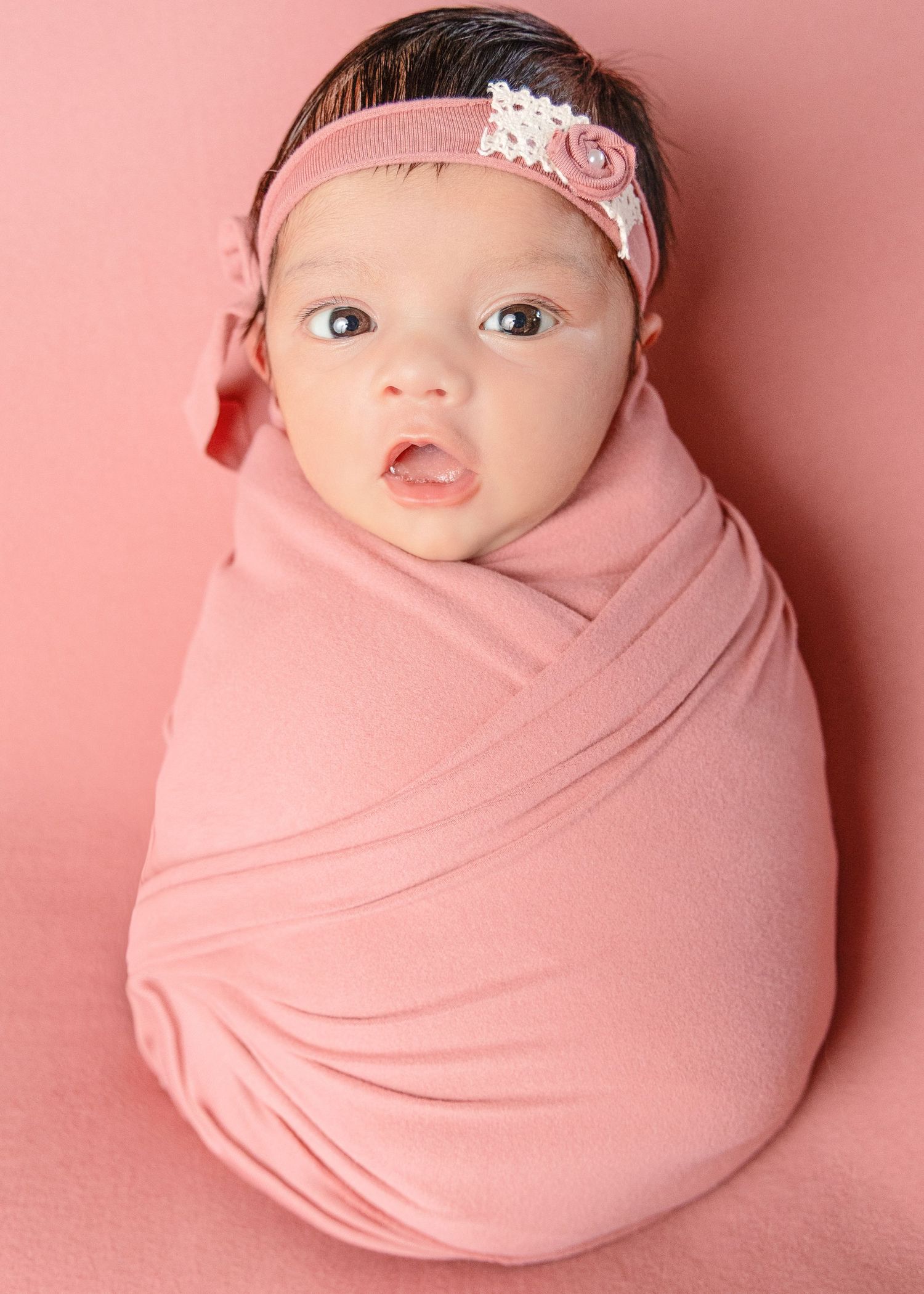 Newborn baby girl swaddled in a pink wrap with a floral headband, posed on a pink background.