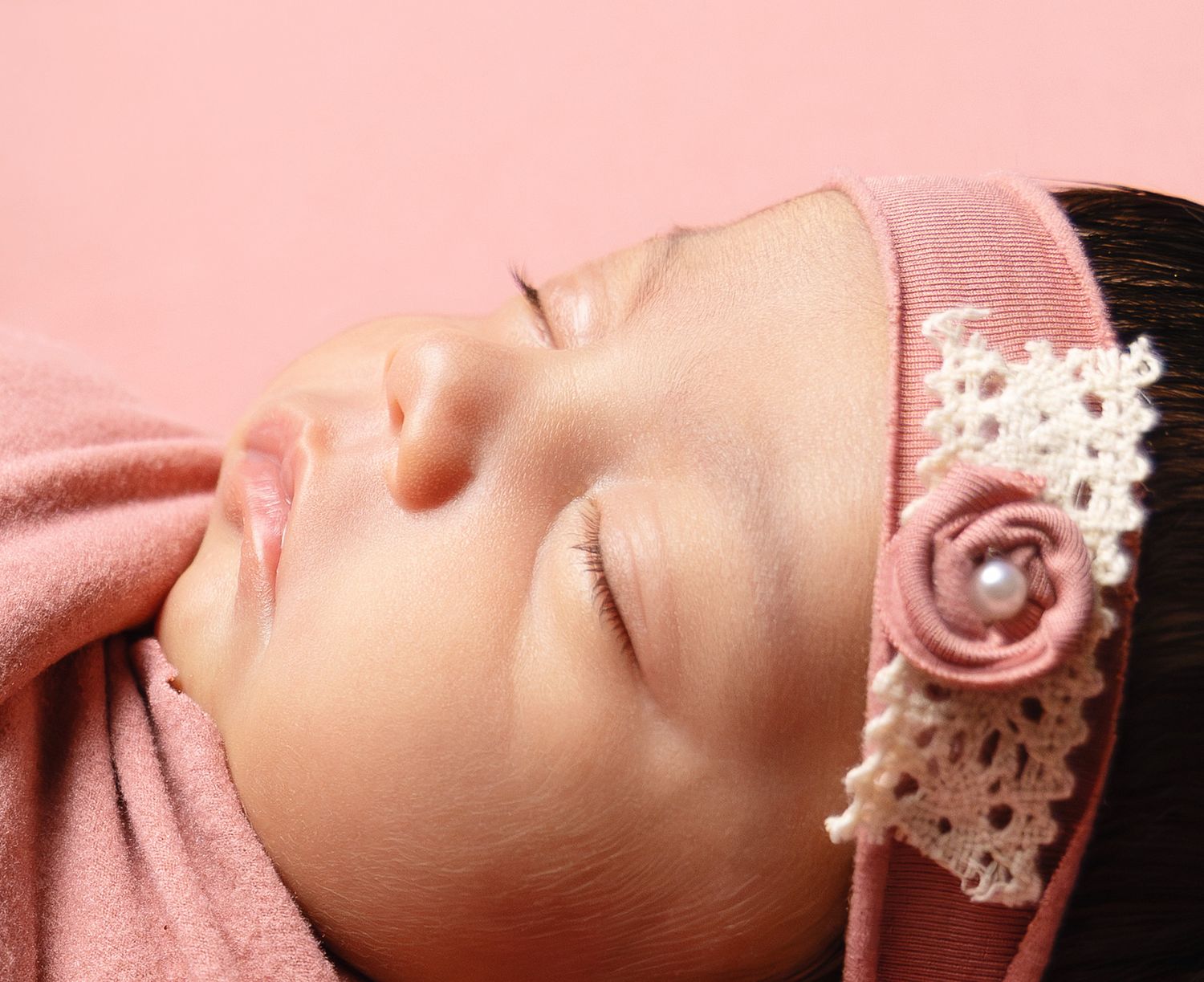 Close-up of sleeping newborn baby with pink headband featuring lace rose on pink background.