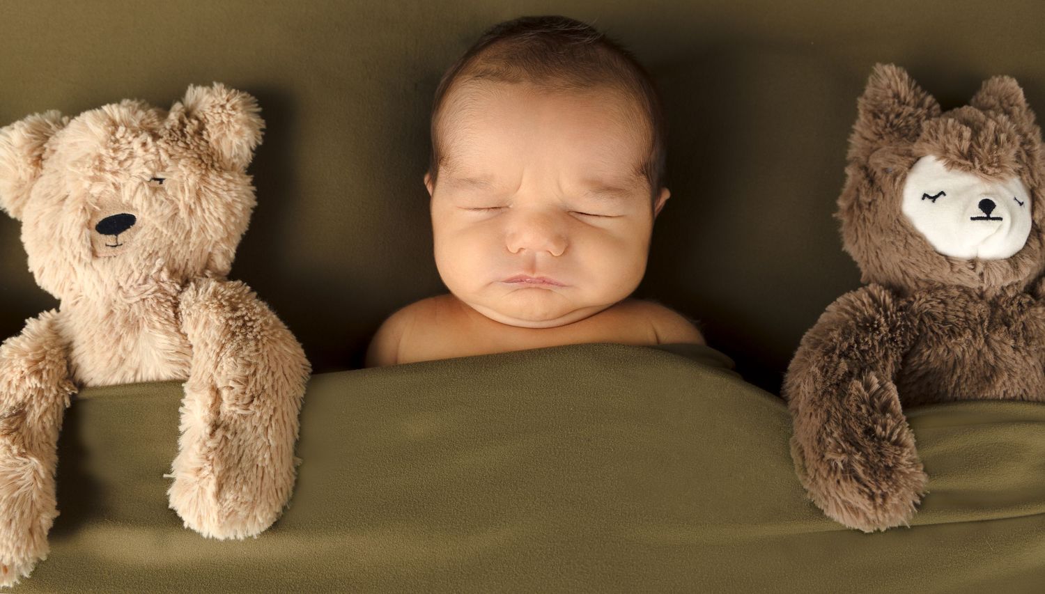 Peaceful newborn baby sleeping tucked under a green blanket between two stuffed teddy bear toys. Artistic newborn photography by Greatest Gi