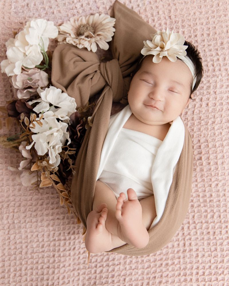 Smiling newborn baby girl wrapped in tan swaddle with white floral headband, posed on pink blanket with cream flowers.