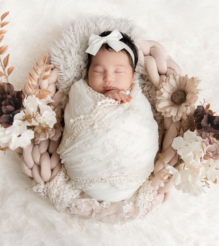 Sleeping newborn girl swaddled in white lace wrap with bow headband in floral boho newborn photography nest.