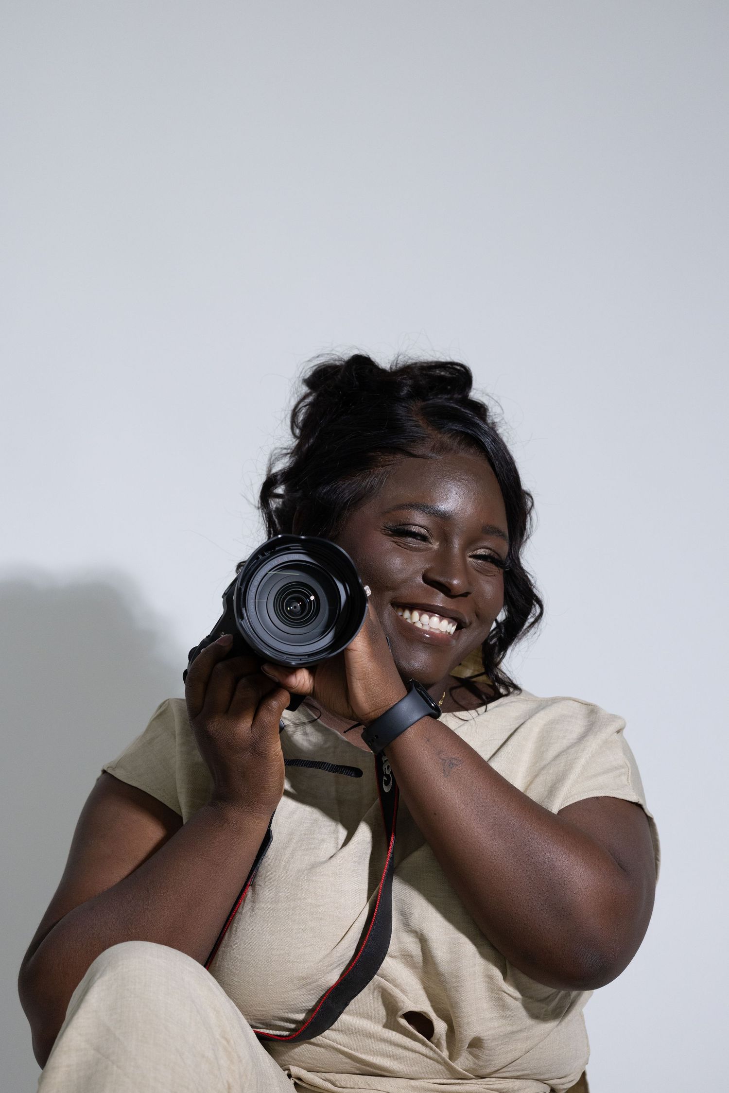 Smiling Black female photographer holding DSLR camera against light gray background.