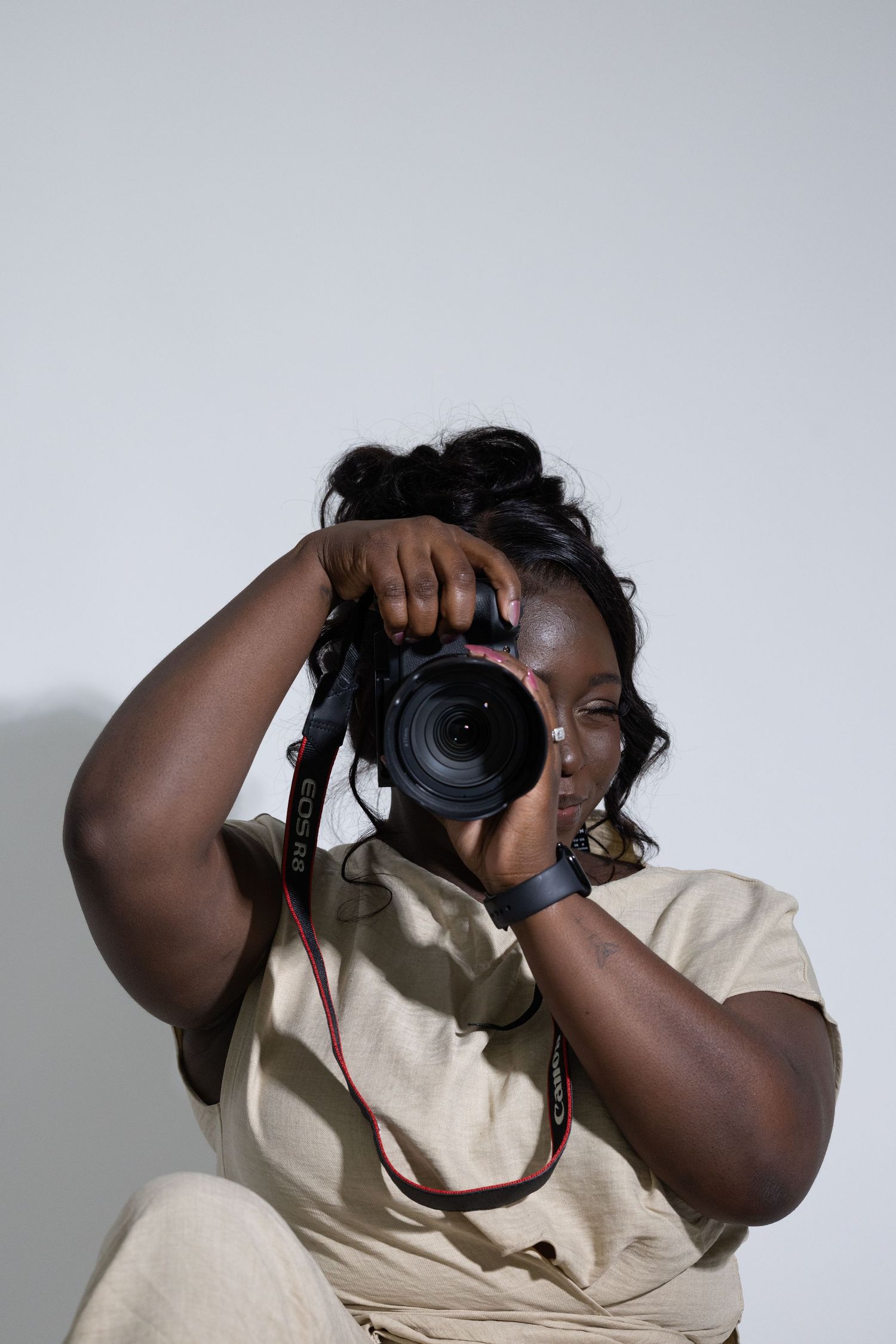 Black female photographer aiming DSLR Canon camera against light gray background.