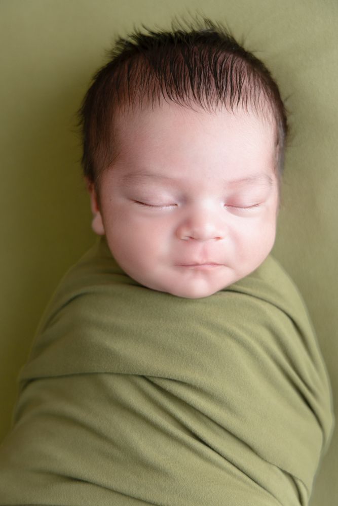 Peaceful newborn baby swaddled in a green wrap, sleeping soundly on a matching green background.