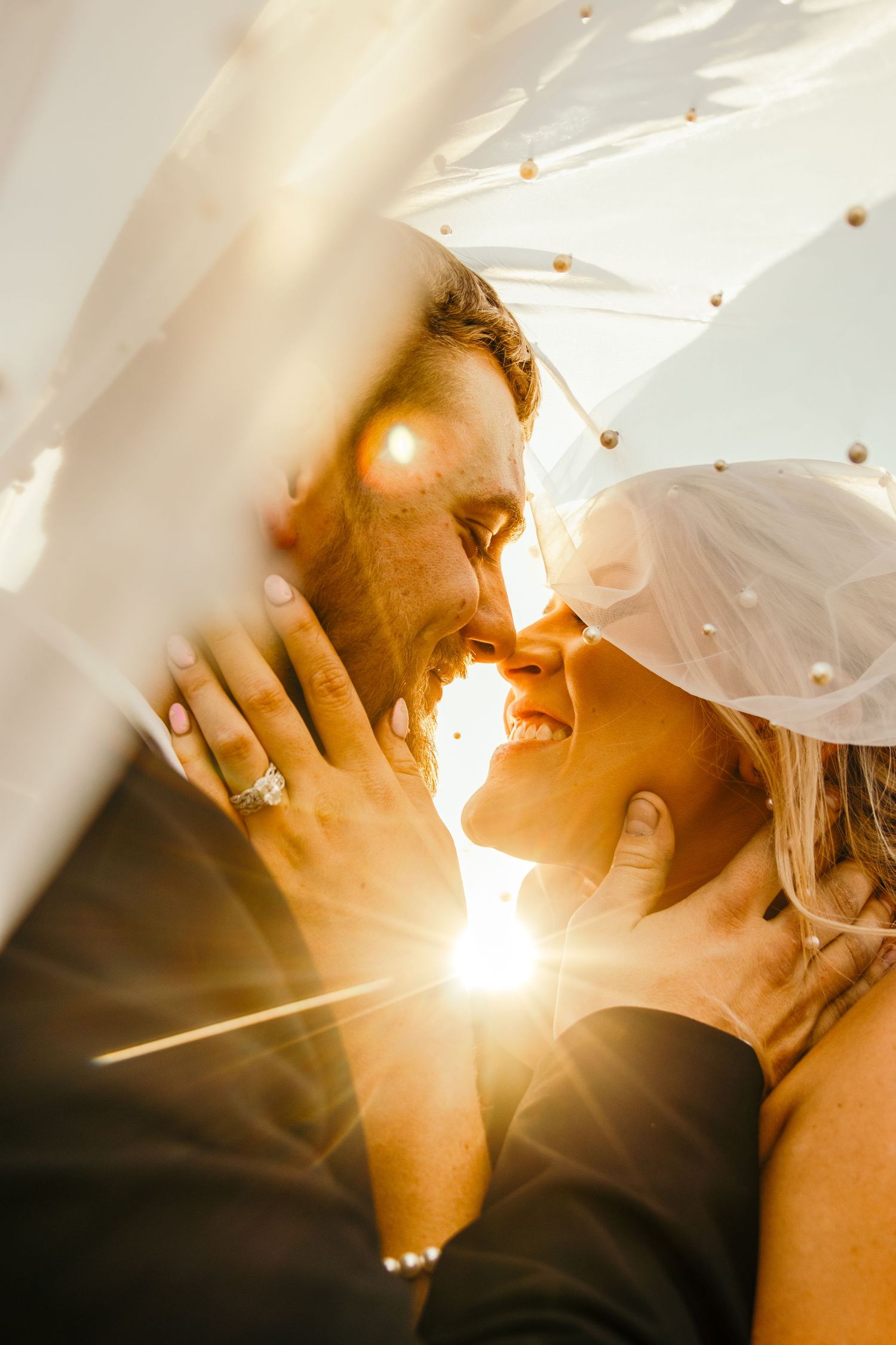 bride and groom nose to nose under veil with sunrays coming through the veil