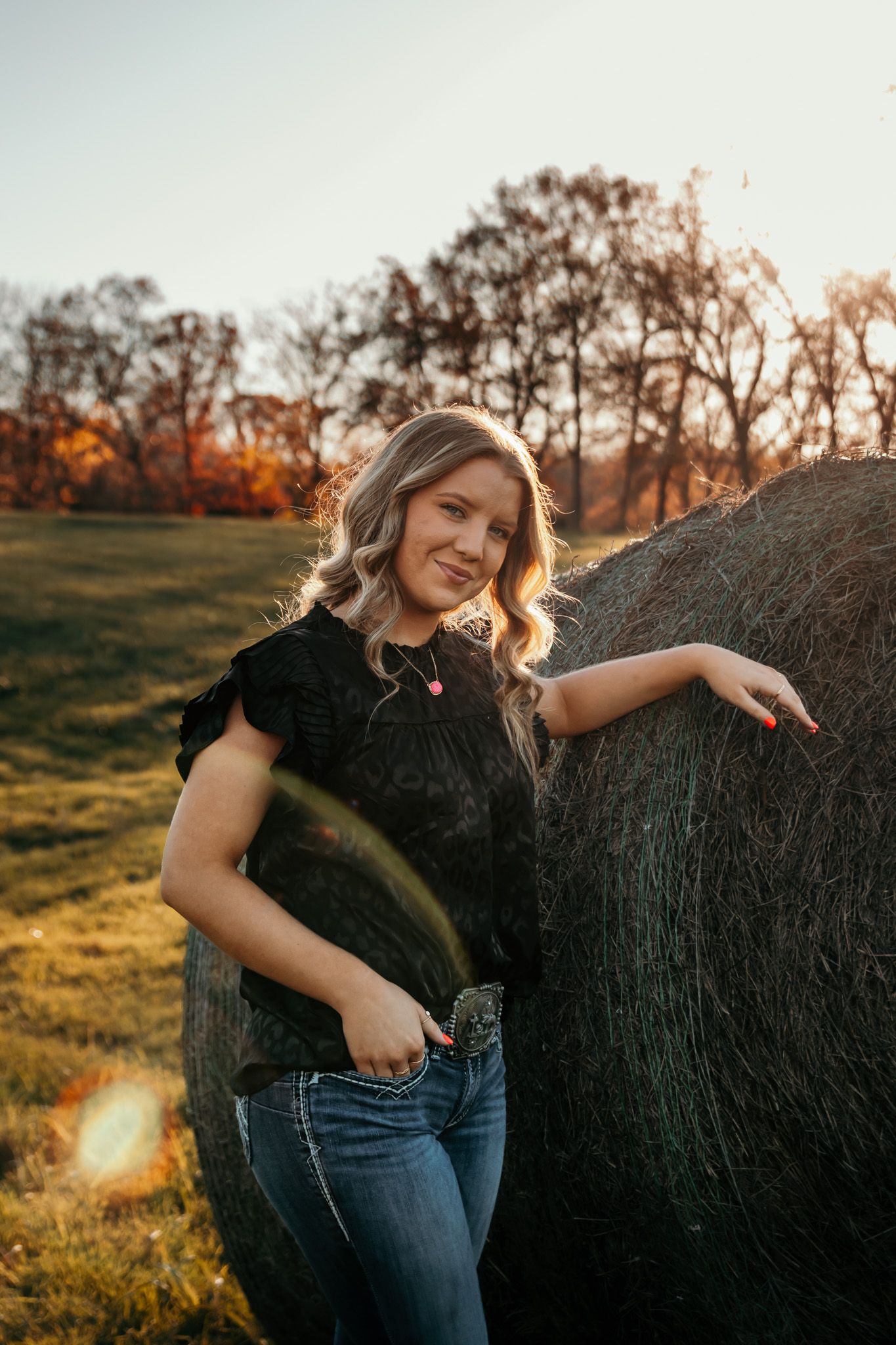 Girl leaning against a haybale while the warm golden sun peeks over hitting her on the face.