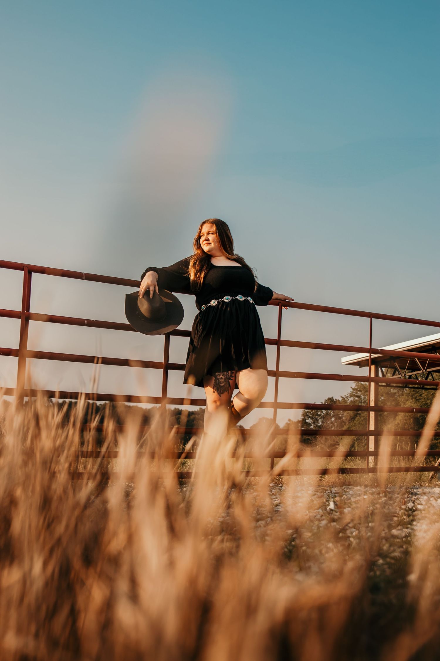 Girl dresses in western attire leaned against a gate while holding her cowboy hat looking off into the distance longingly.