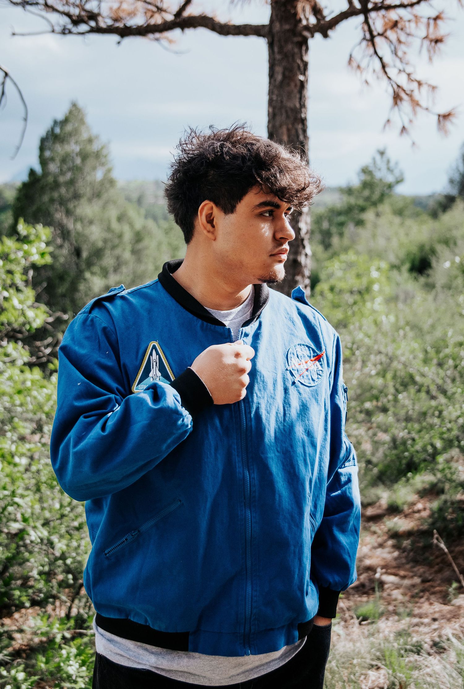 High school senior wearing a blue NASA jacket poses against pine trees in an outdoor setting at Palmer Park in Colorado Springs
