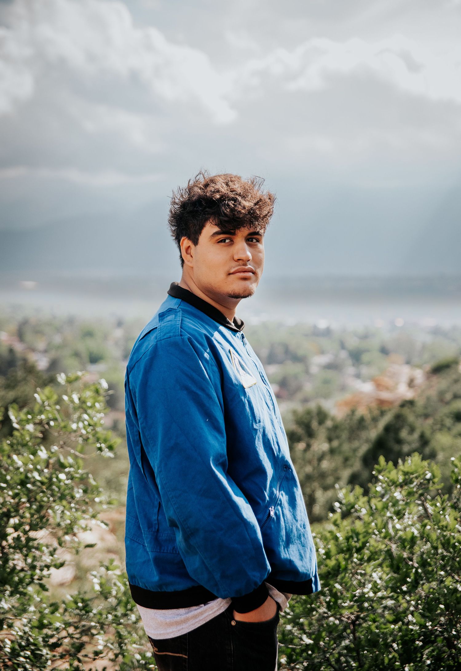 High school senior in blue NASA uniform stands among bushes on hilltop overlooking valley at Palmer Park in Colorado Springs