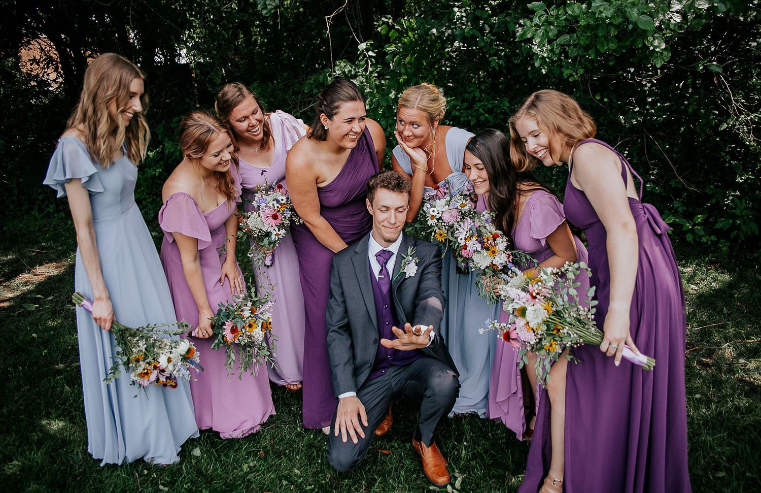 Playful photo of bridal party lifting bride's veil in outdoor setting.