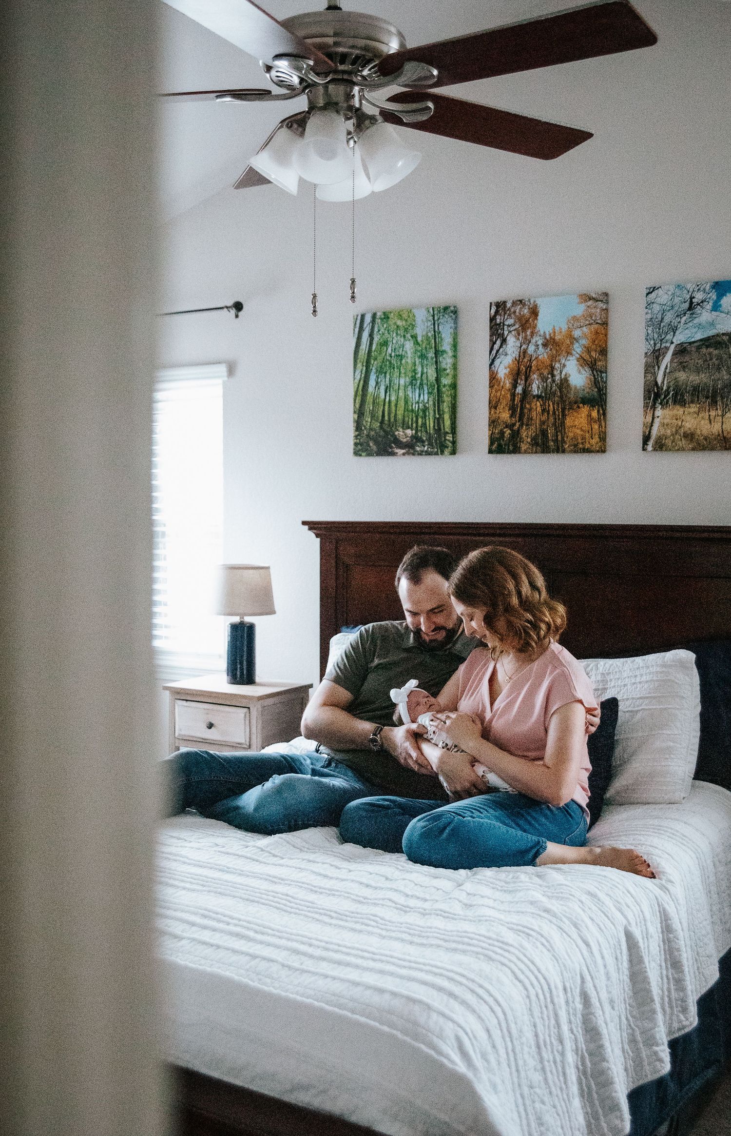 Couple sitting on bed with forest artwork and ceiling fan above while holding baby in cozy bedroom setting.