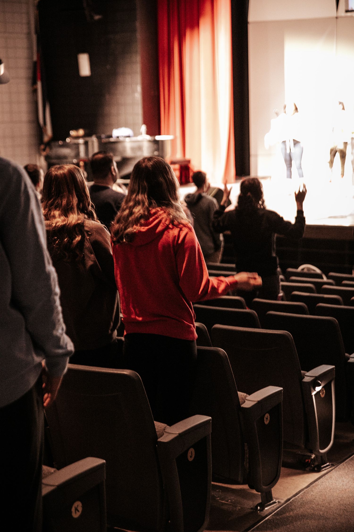 Audience members standing in theater seats during a performance with red curtain visible.