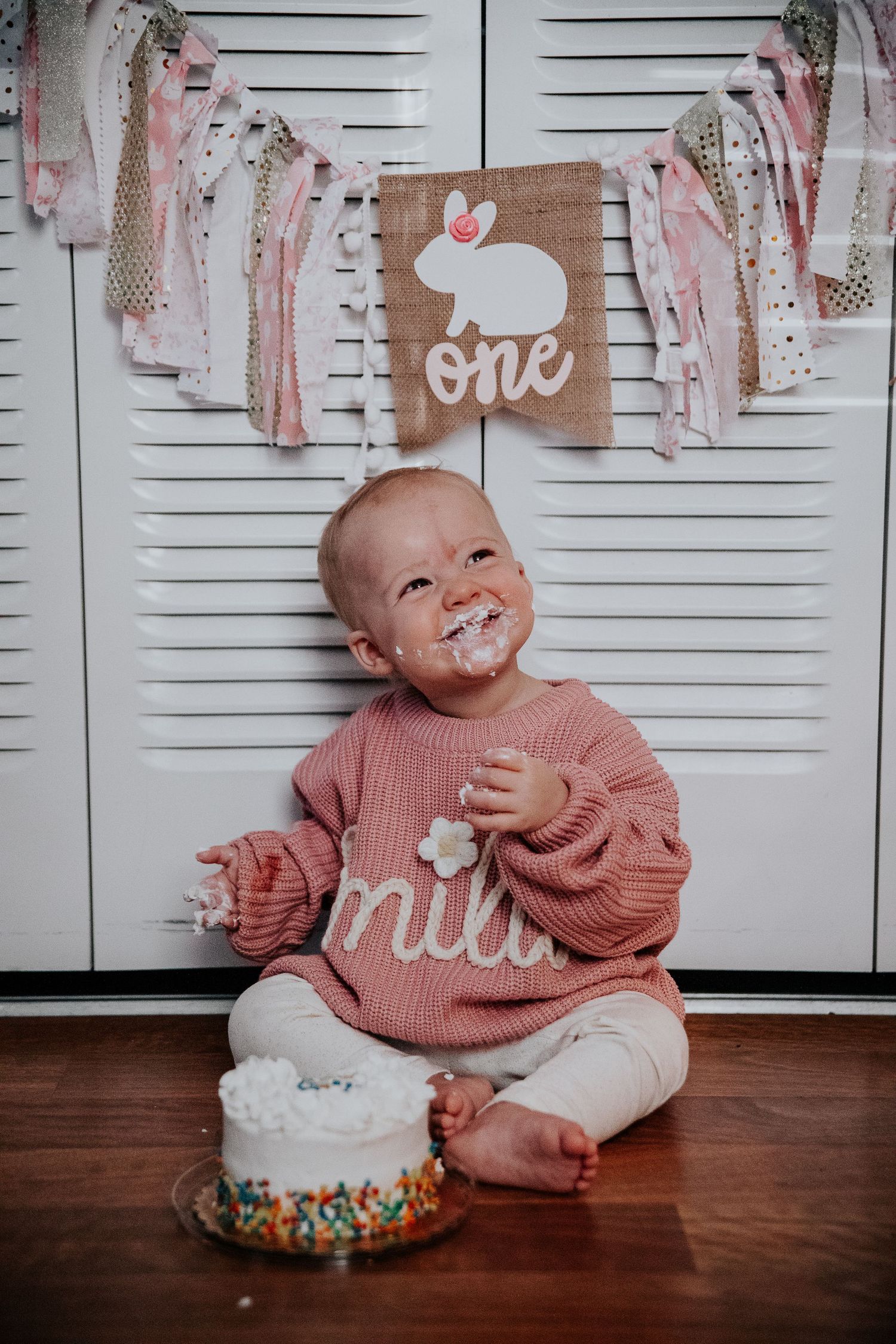 A first birthday celebration with pink decorations and a smash cake featuring a bunny banner and pink sweater.