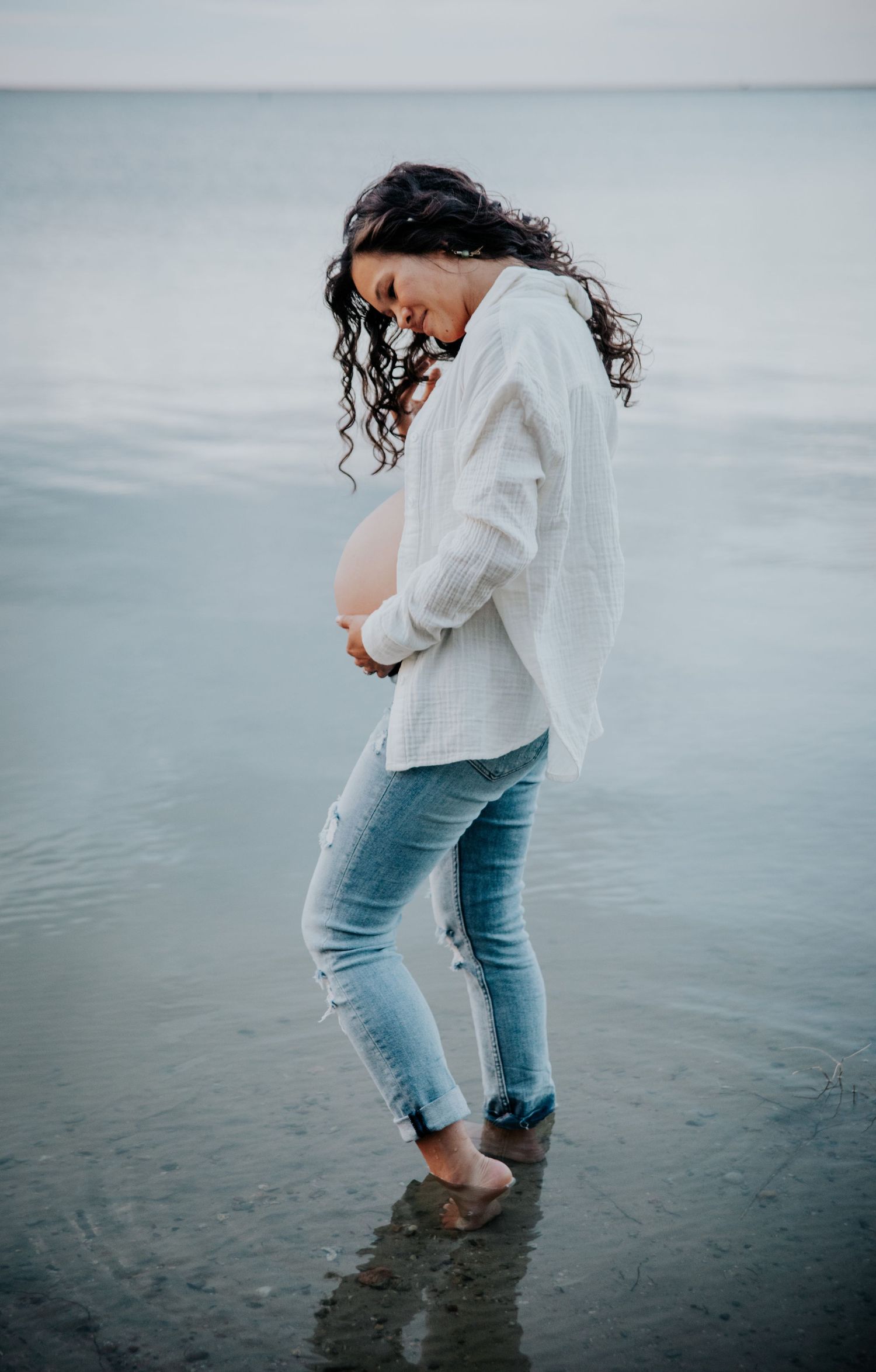 Woman in distressed jeans and white sweater wading in calm waters at sunset.