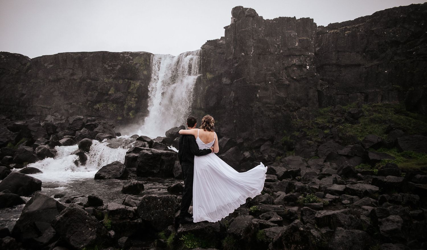 A flowing white dress catches the wind as a couple embraces near a majestic waterfall.