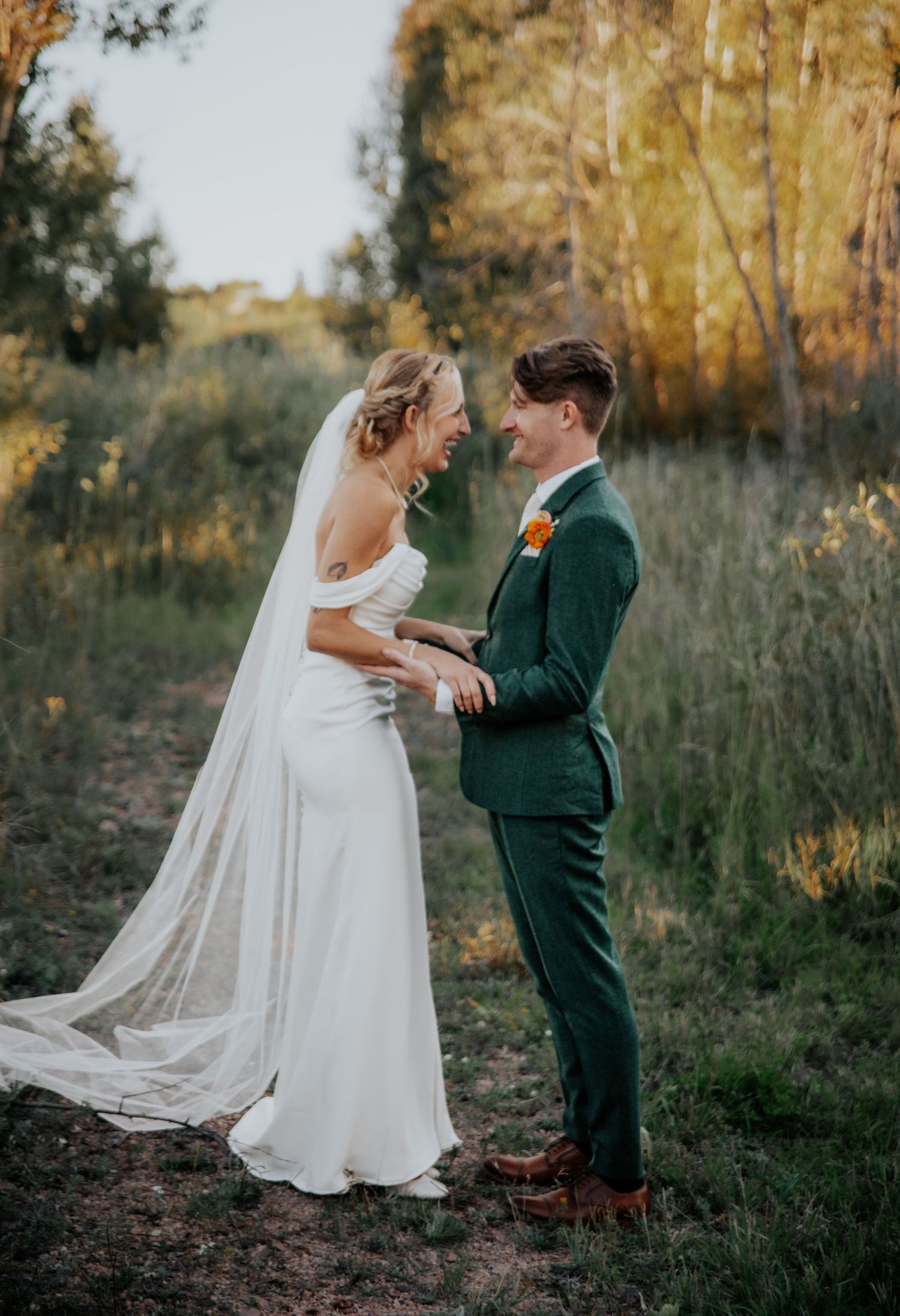 Elegant wedding couple embracing in meadow with flowing veil and forest backdrop.