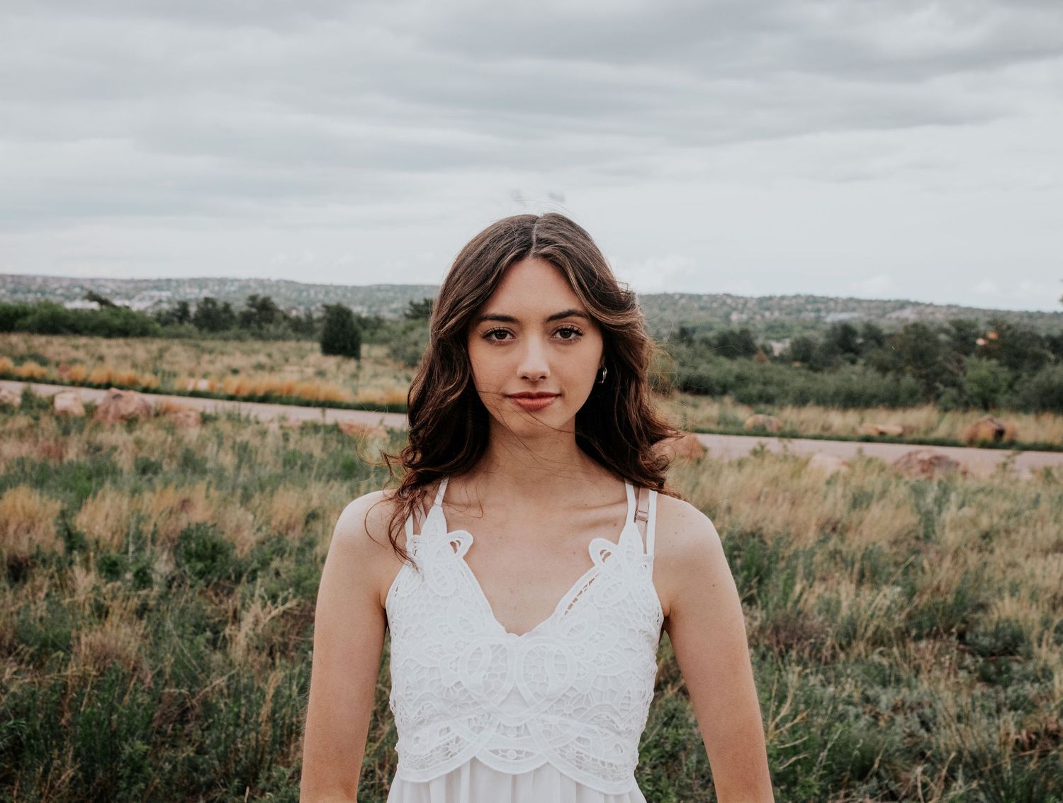 A figure in white lace poses gracefully in an open field under cloudy skies.