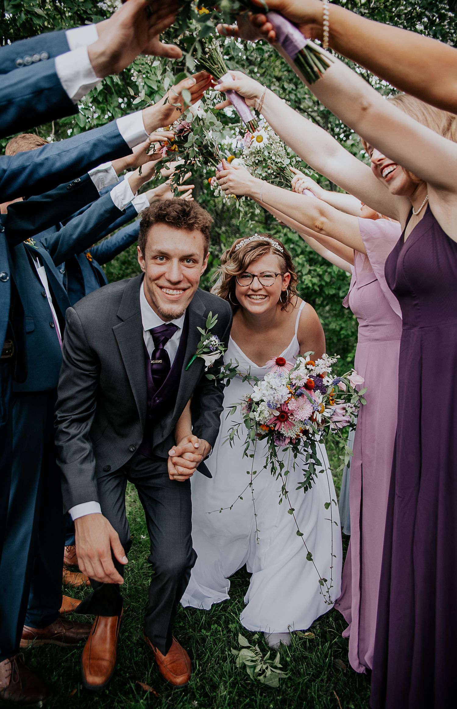 Wedding party forms tunnel with raised arms over couple in outdoor celebration.