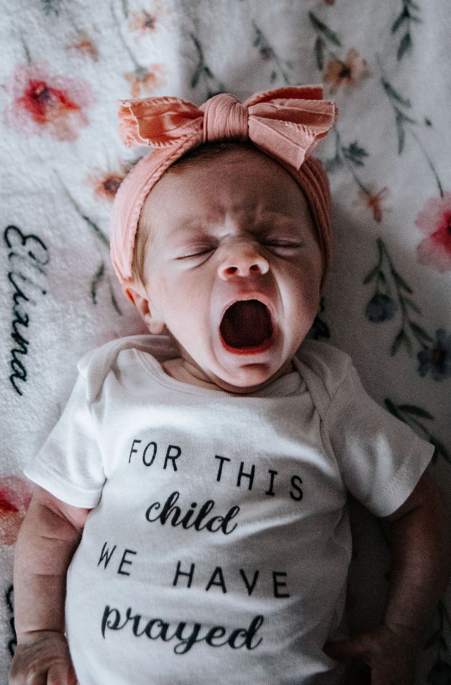 A baby in a white onesie with text and pink bow headband lying on floral patterned fabric.