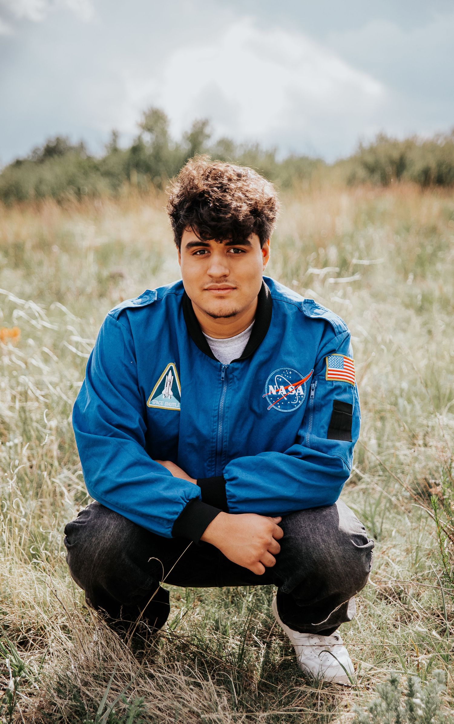 High school senior wearing NASA flight jacket poses in tall grass field under cloudy skies at Palmer Park in Colorado Springs