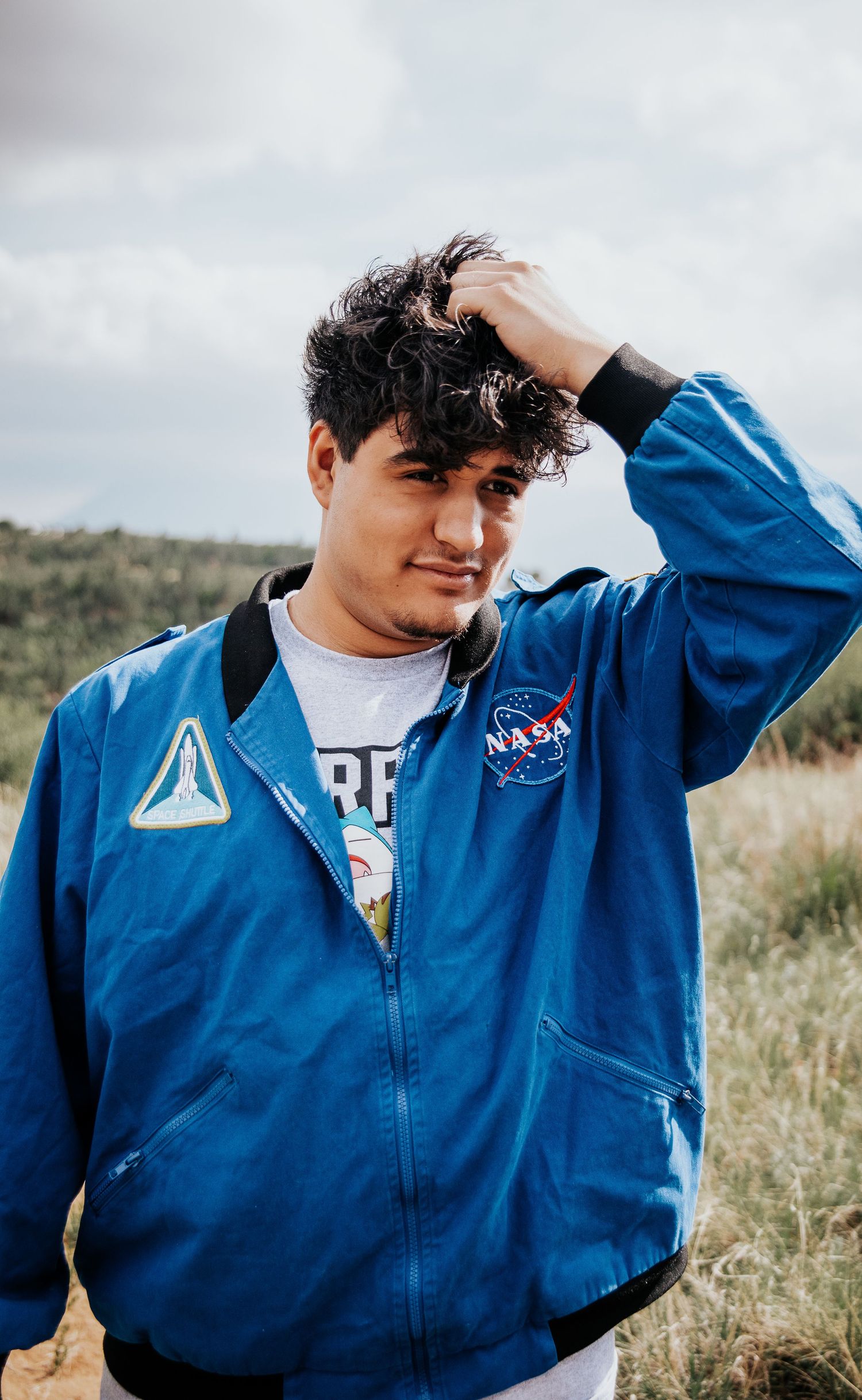 High school senior in blue NASA jacket poses outdoors with hand in hair against natural landscape at Palmer Park in Colorado Springs