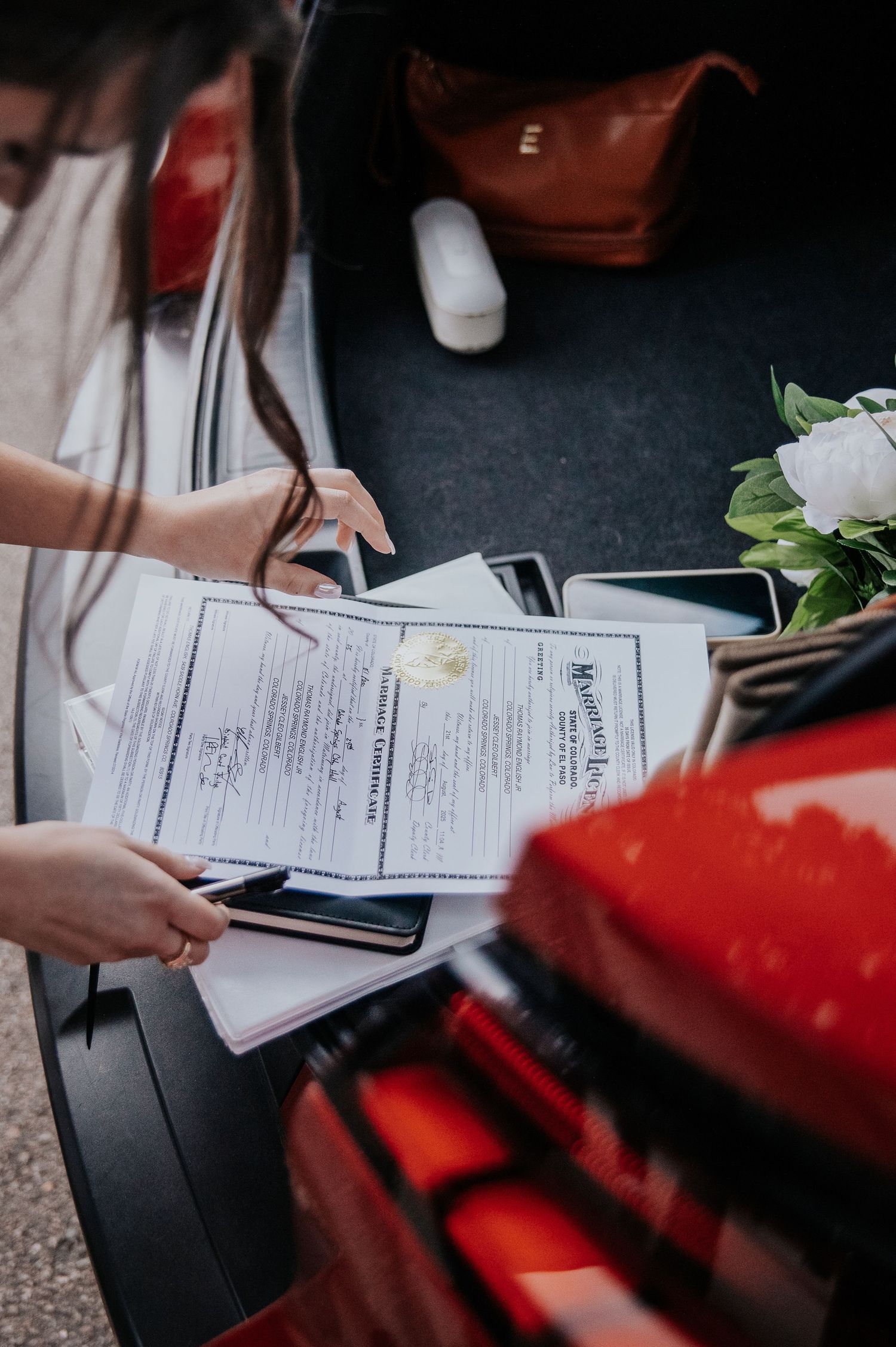 Wedding paperwork and documents being signed on a dark surface with flowers visible.
