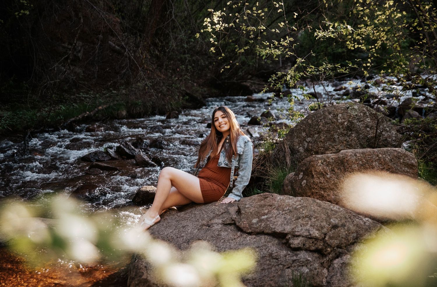 Casual portrait on rocky terrain by stream wearing denim jacket and brown dress.