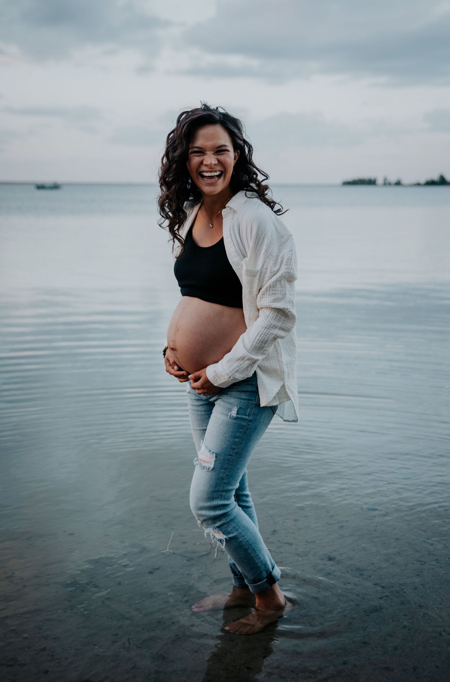 Expecting mother laughs joyfully while standing at the beach in casual attire.