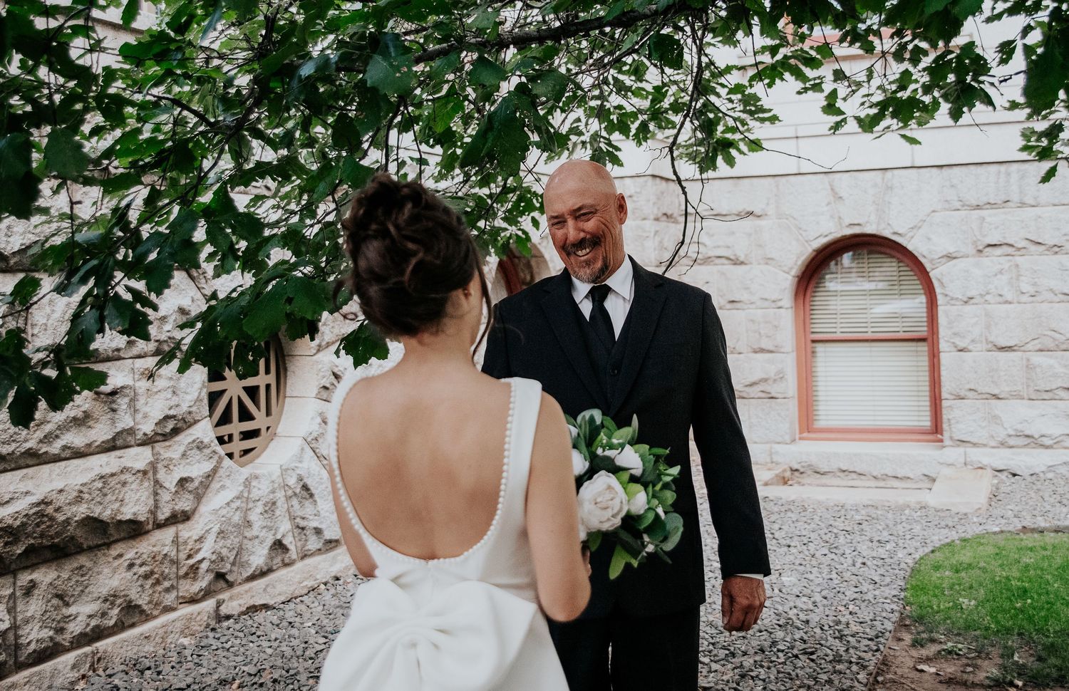 A couple shares a tender moment during their wedding ceremony outside a stone building with arched windows.