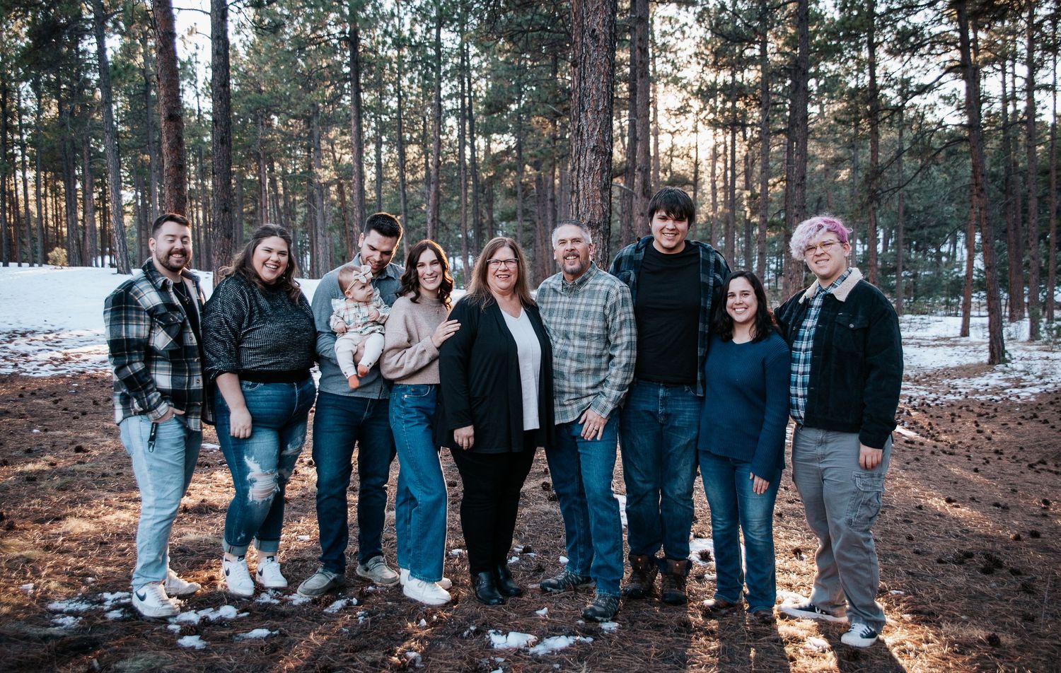 Friends and family gather outdoors for a group photo in a pine forest setting.