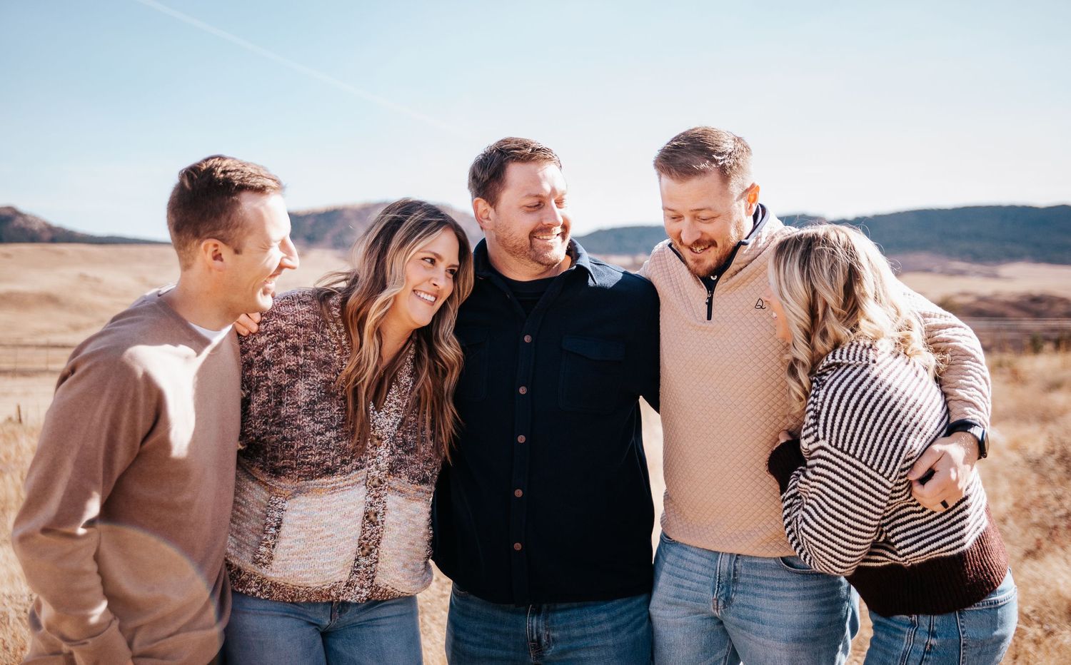 A group in casual attire stands together embracing outdoors against a scenic natural backdrop during golden hour.