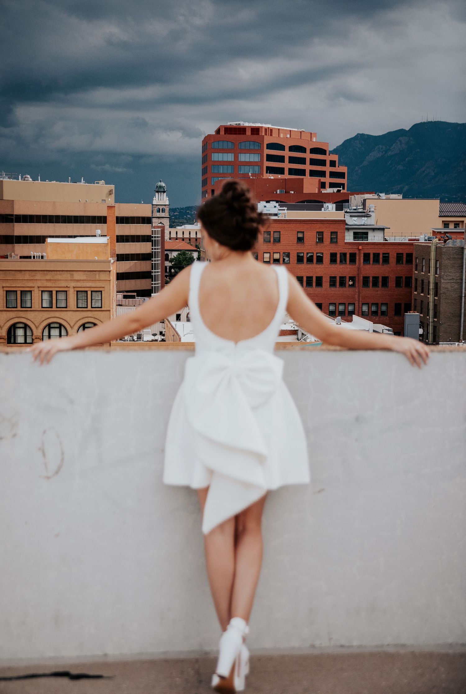 A bride in a white dress poses dramatically on a rooftop with urban buildings and mountains in the background.