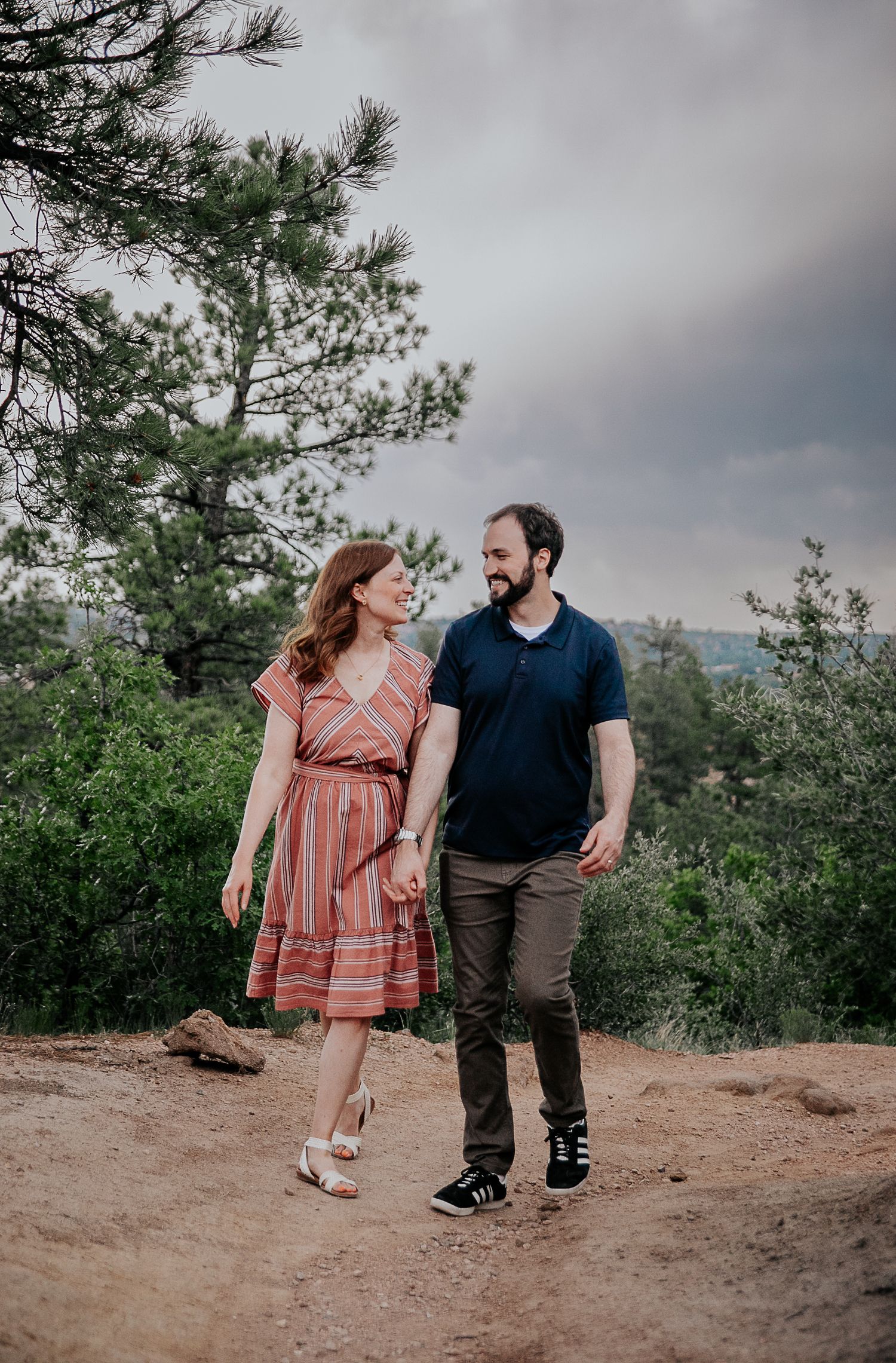 A couple walks together on a dirt path surrounded by pine trees and greenery.
