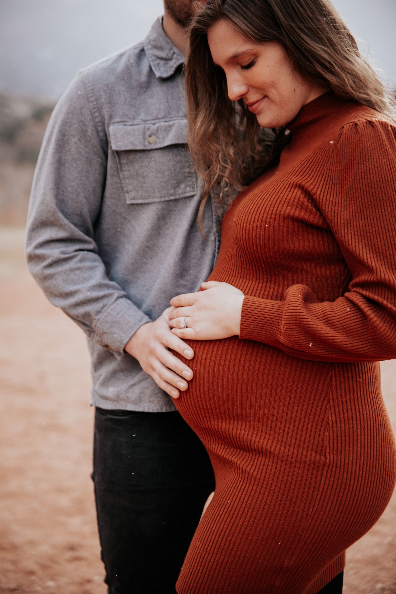 A person in a rust-colored sweater shows their baby bump while being held from behind.