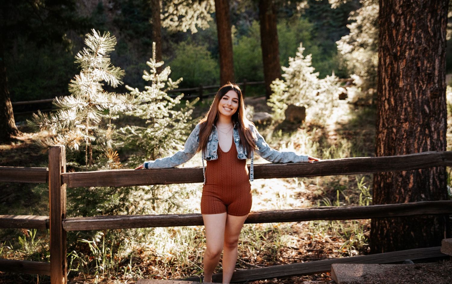 Standing against rustic wooden fence in forest with illuminated trees in background.