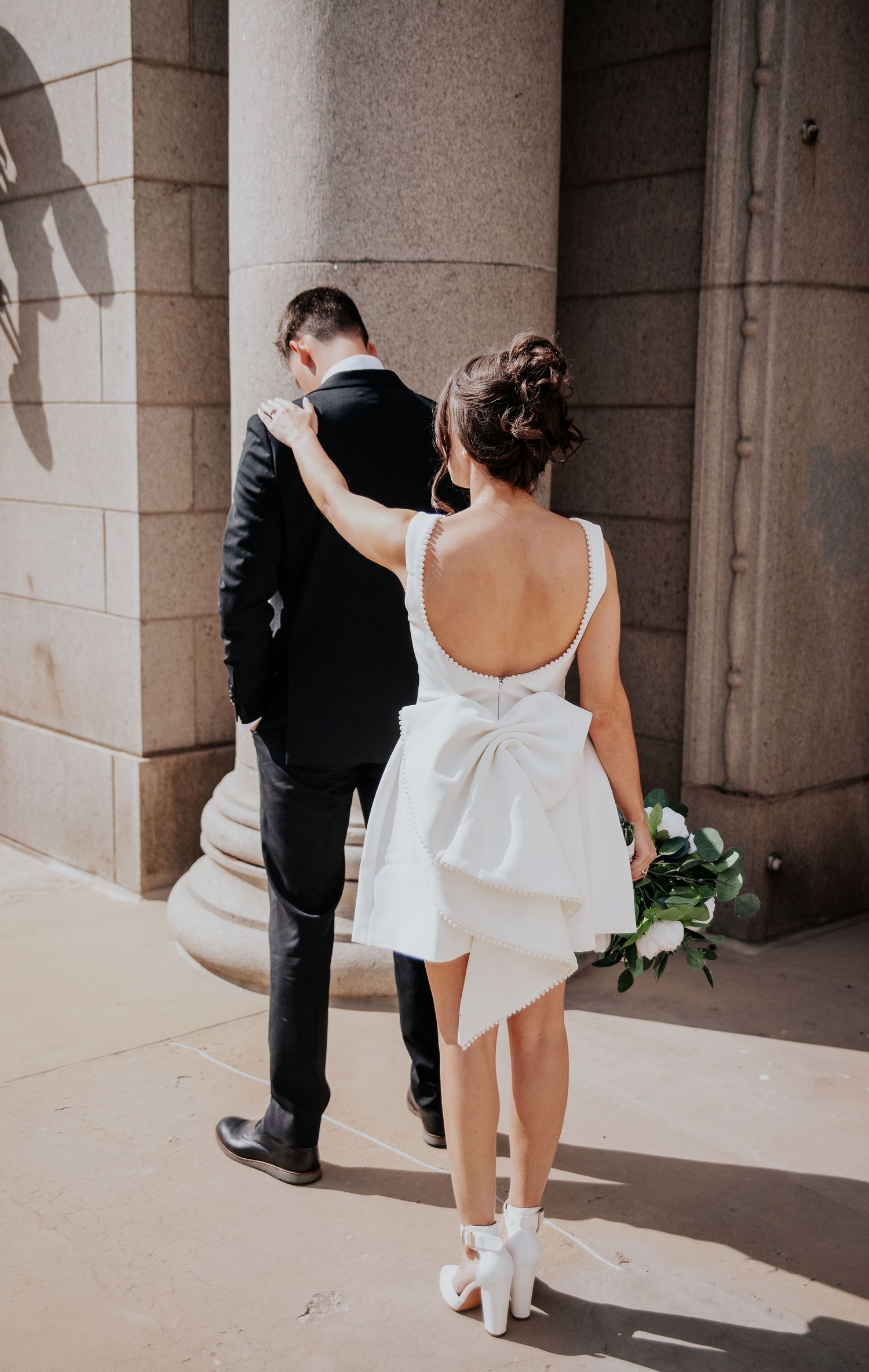 A couple in wedding attire walks through a columned archway casting dramatic shadows on the stone floor.