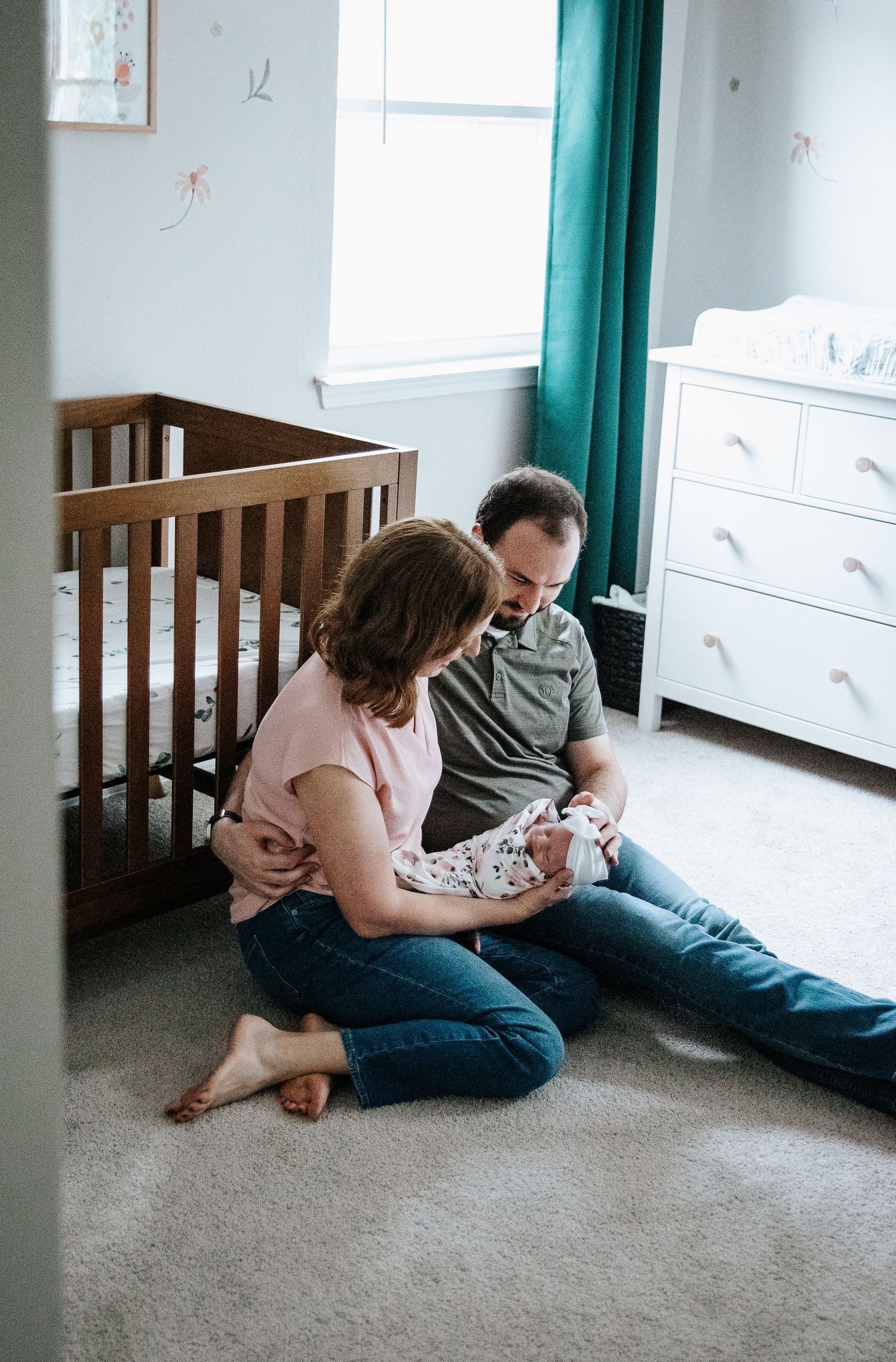 Parents sitting on nursery floor beside wooden crib and white dresser with teal curtains in background.
