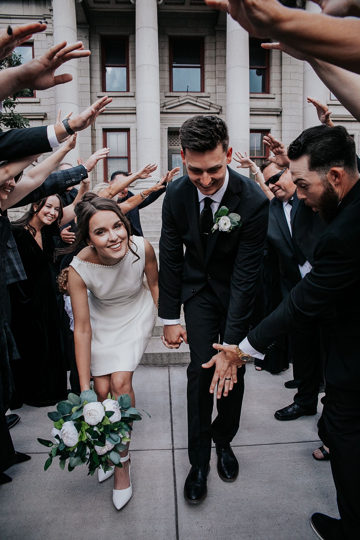 Wedding party and guests celebrating with raised hands outside historic building.
